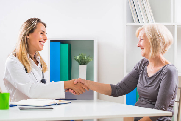 Female doctor and happy senior woman patient shaking hands in doctor's office. - Foto, Bild