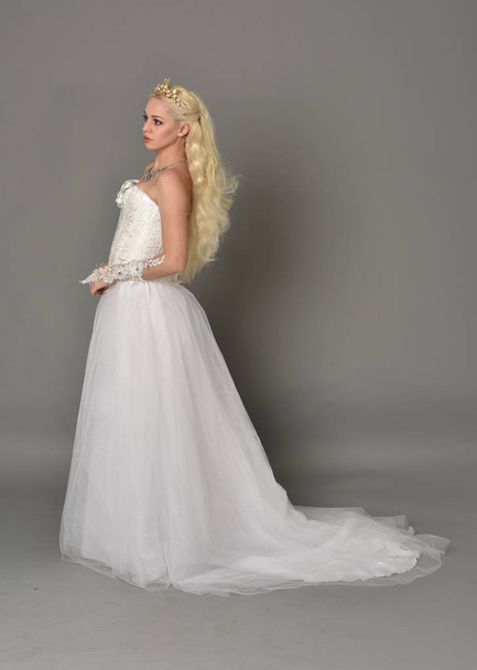 full length portrait of blonde  girl wearing white fantasy gown. standing pose in side profile, grey studio background. - Фото, изображение