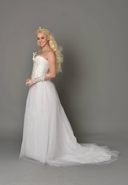 full length portrait of blonde  girl wearing white fantasy gown. standing pose in side profile, grey studio background. - Foto, afbeelding