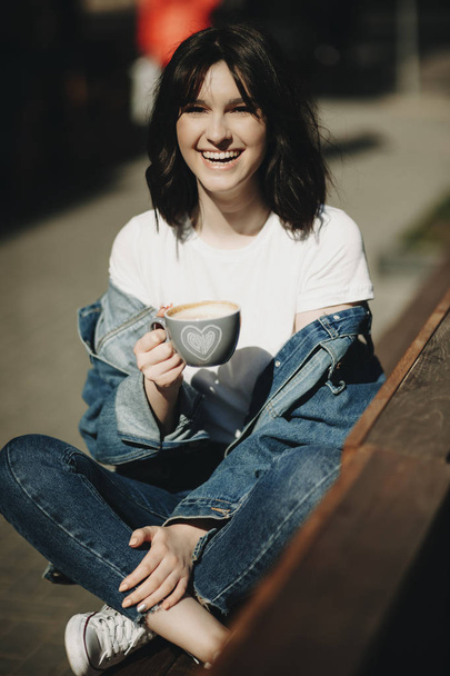 Laughing female with dark hair in simple jeans clothes sitting with crossed legs on wooden bench outdoors and holding cup with drink in hand looking at camera on blurred background - Zdjęcie, obraz