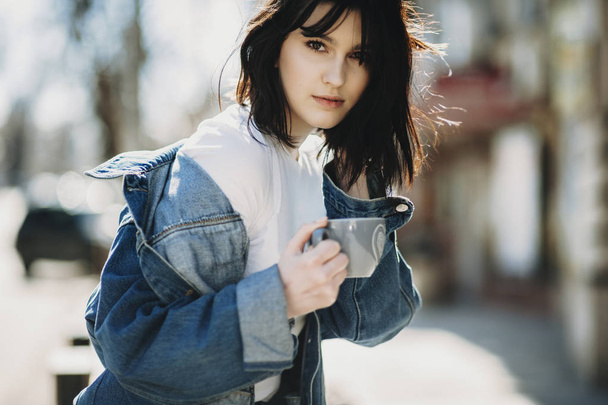 Crop side view of trendy young brunette female holding cup and touching hair standing leaning forward and looking at camera on blurred street background - Foto, immagini