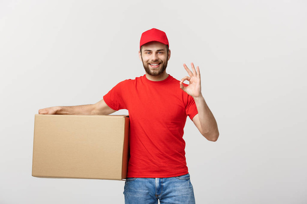 Allegro giovane uomo di consegna con cappuccio rosso in piedi con pacco casella postale isolato su sfondo bianco mostrando gesto ok. - Foto, immagini