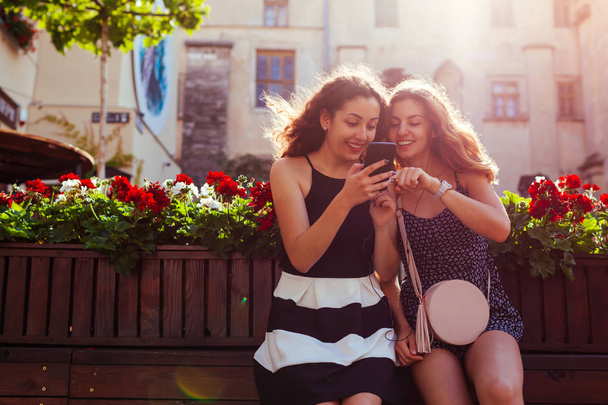 Joyeuses amies vérifiant les réseaux sociaux en utilisant le téléphone par café dans la rue de la ville. Loisirs d'été. Les étudiants s'amusent
 - Photo, image