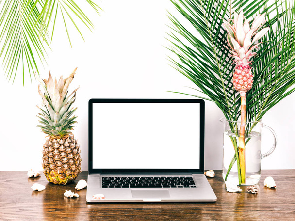 Laptop com uma tela branca em branco. Computador desktop whith freelancer, ramos de palma e abacaxi rosa decorativo em um vaso em uma mesa de madeira
. - Foto, Imagem