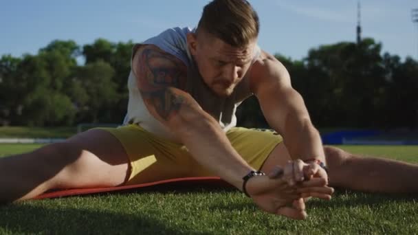 Desportista muscular tatuado sentado no tapete e se curvando para frente para esticar os músculos da perna na grama verde do campo atlético
 - Filmagem, Vídeo