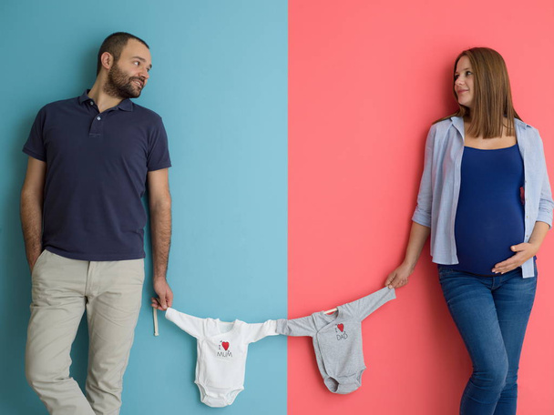 Beautiful pregnant woman and her husband expecting baby holding baby bodysuits and smiling over colorful background - Fotoğraf, Görsel