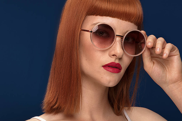 Portrait of beautiful redhead young woman wearing fashionable sunglasses. Girl in glamour makeup. Haircut with fringe. Blue background. - Foto, imagen