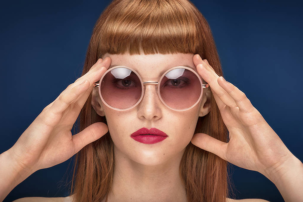 Portrait of beautiful redhead young woman wearing fashionable sunglasses. Girl in glamour makeup. Haircut with fringe. Blue background. - Foto, Imagem
