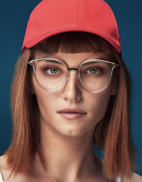 Portrait of attractive young redhead woman with fashionable eyeglasses and cap. Girl looking at camera. Blue background. - Photo, Image