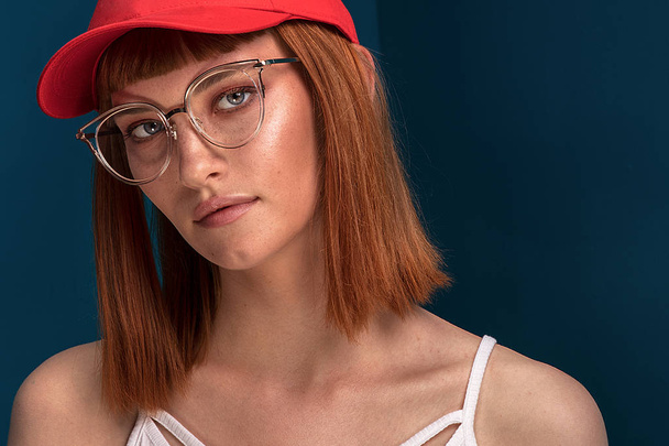 Portrait of attractive young redhead woman with fashionable eyeglasses and cap. Girl looking at camera. Blue background. - Φωτογραφία, εικόνα