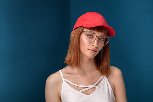 Portrait of attractive young redhead woman with fashionable eyeglasses and cap. Girl looking at camera. Blue background. - Фото, зображення