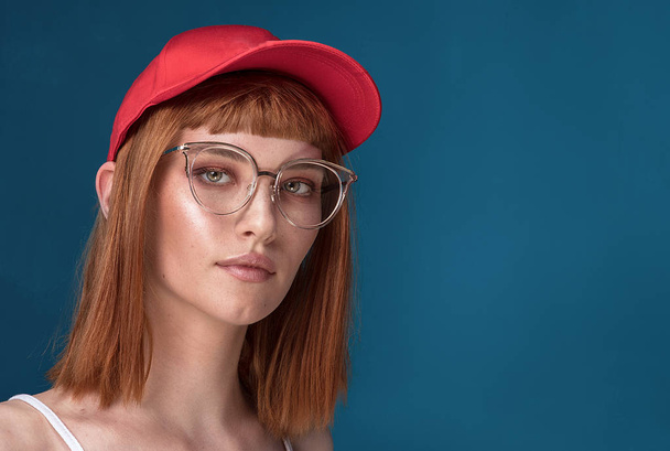 Portrait of attractive young redhead woman with fashionable eyeglasses and cap. Girl looking at camera. Blue background. - Photo, image