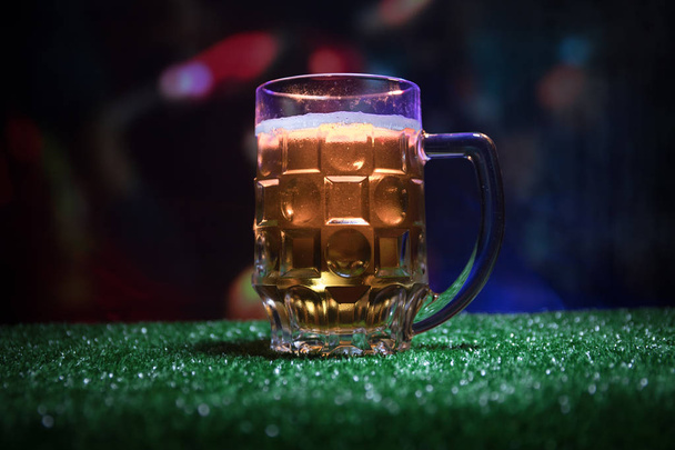 Creative concept. Pint of fresh beer on grass with blurred stadium on background with fog and light. Glass of lager beer ready for drink. Copy space. Selective focus - Photo, Image