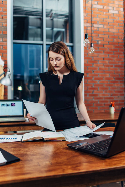Serious woman reading papers studying resumes standing at work desk in stylish office - Foto, Bild