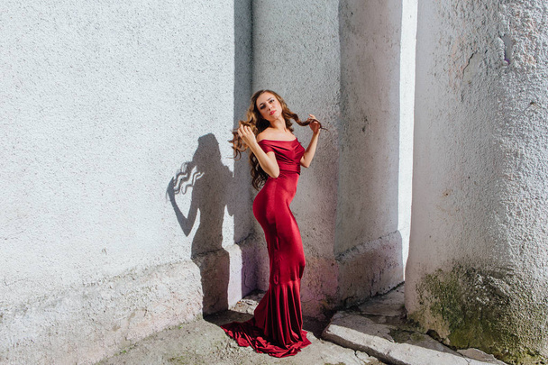 Beautiful young woman in a long red evening gown with a train standing next to the old wall - Foto, Imagem