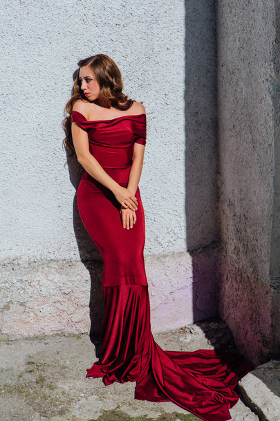 Beautiful young woman in a long red evening gown with a train standing next to the old wall - Foto, Bild