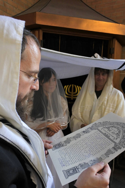Rabbiner liest die Ketuba (jüdischer Hochzeitsvertrag) einer jüdischen Braut und eines Bräutigams unter einer Chupa (Baldachin aus Tallit, der ein jüdisches Haus darstellt) in einer Synagoge a am Hochzeitstag nach ihrer Konvertierung zum Judentum. - Foto, Bild