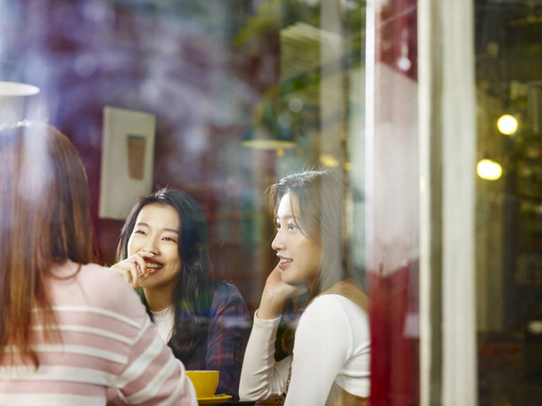 tres feliz hermosa joven asiático mujeres sentado a la mesa charlando hablando en la cafetería o casa de té, disparado a través de cristal de la ventana
. - Foto, imagen