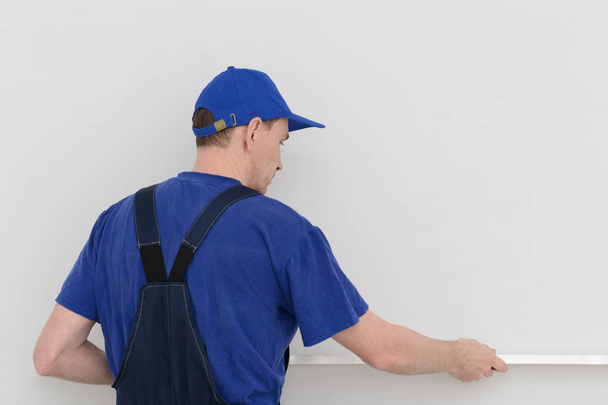 εργαζόμενος σε μπλε στολή μετρά την καμπυλότητα του τοίχου με ένα επίπεδο - Φωτογραφία, εικόνα