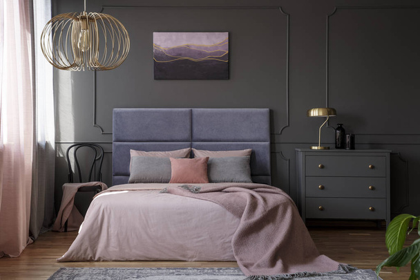 Gold lamp on grey cabinet next to pink bed in elegant pastel bedroom interior with chair - Foto, Imagen