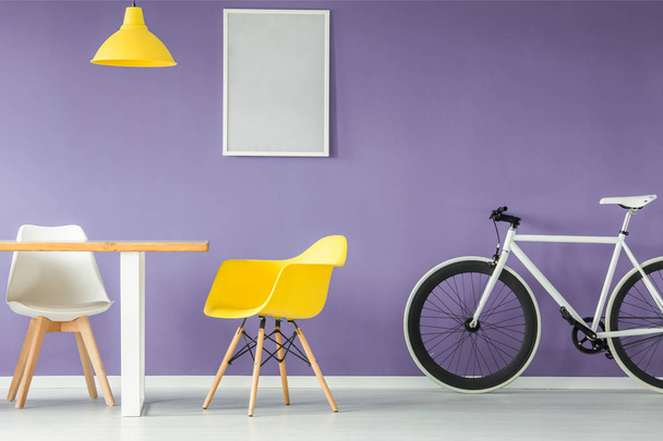 Minimal, modern interior with a white and yellow chair, a bicycle, an empty table and a hanging yellow lamp against purple background wall with a mockup poster - Foto, Bild