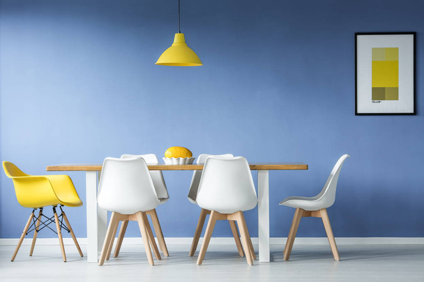 Minimal, modern dining room in interior with a table and contrasting white and yellow chairs around it, against blue wall with a mockup poster - Фото, зображення