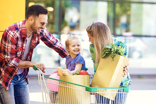 Young smiling parents and their cute daughter with shopping cart full of fresh food having fun in front of hypermarket. - Fotoğraf, Görsel