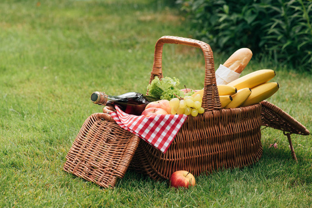 tasty fruits and bottle of champagne in wicker basket on green grass at picnic - Photo, Image