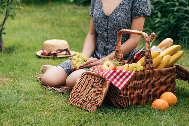 cropped image of woman sitting on green grass at picnic and holding grapes - Photo, Image
