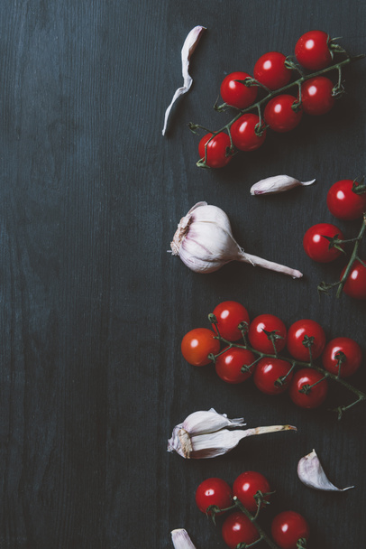 top view of red cherry tomatoes and garlic on black wooden background - Fotoğraf, Görsel