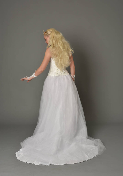 Ganzkörperporträt eines blonden Mädchens im weißen Kleid, das mit dem Rücken zur Kamera posiert. grauer Studiohintergrund. - Foto, Bild