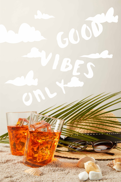 Close-up van stro hoed, cocktails, schelpen, zonnebril en palmtak op zand op grijze achtergrond met "good vibes alleen" teken - Foto, afbeelding