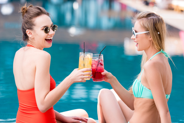 side view of attractive young women clinking glasses of cocktails at poolside - Photo, Image