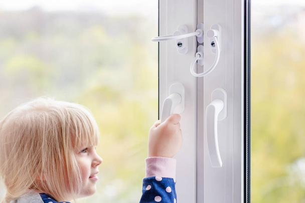 Little cute toddler girl trying to open window in apartment at high-tower building. Children window protection lock. Cable safety guard prevent opening window by child. Prevention of falling accident. - Photo, Image
