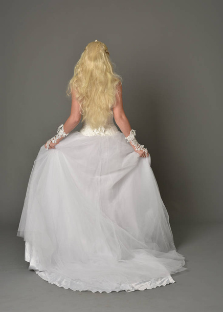 full length portrait of blonde girl wearing white gown, standing pose with back to the camera. grey studio background. - Фото, зображення