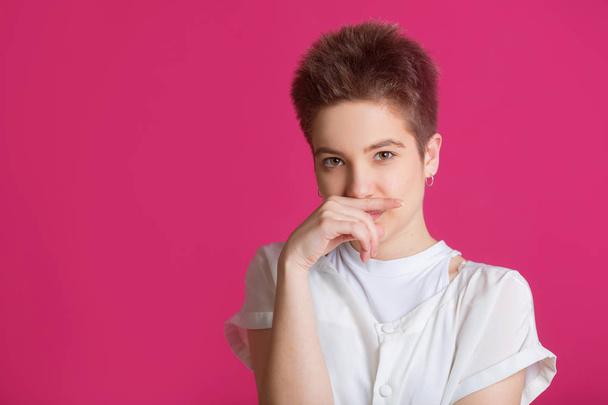 beautiful young girl with short hairdo in a white T-shirt on a pink background with a displeased face - Zdjęcie, obraz