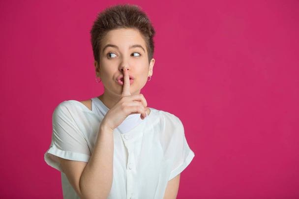 beautiful young girl with short hairdo in a white T-shirt on a pink background with a gesture of silence - Фото, изображение