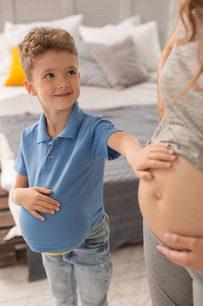 Curly cute son with ball under shirt playing with pregnant mother - Photo, image