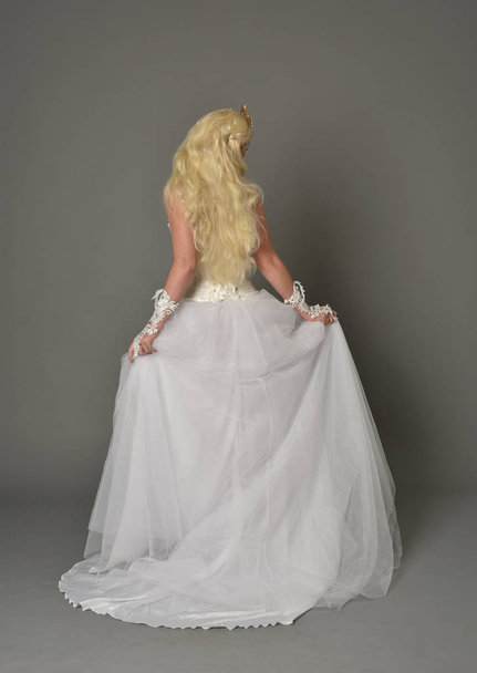 Ganzkörperporträt eines blonden Mädchens im weißen Kleid, das mit dem Rücken zur Kamera posiert. grauer Studiohintergrund. - Foto, Bild