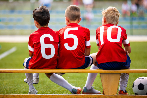 Drużyna dzieci piłka nożna. Dzieci podstaw gracze siedzący na ławce. Turniej sportowy piłki nożnej dla młodych chłopców. Troje dzieci oglądania meczu piłki nożnej na boisku - Zdjęcie, obraz
