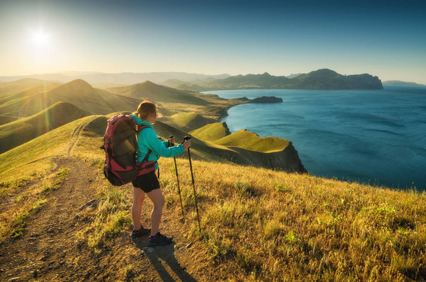 Girl hiker with backpack and trekking sticks standing on a hill in a Crimea mountain valley and enjoy the sunset over the sea. Travel and adventure concept. - Valokuva, kuva