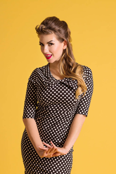 Pretty young woman in dress with hairdo stands in yellow studio, pin up style - Photo, Image