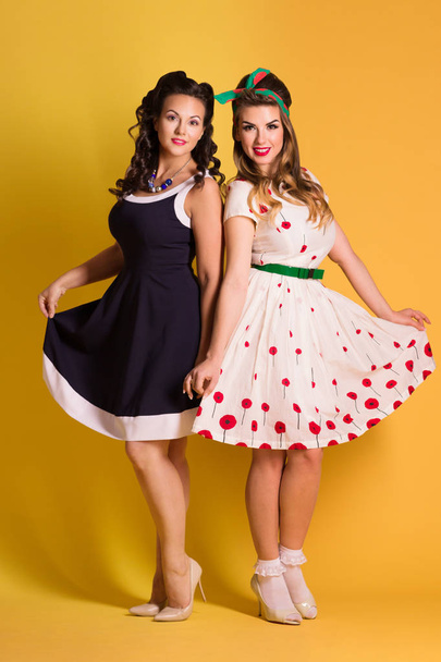 Two pretty women in dresses pose in yellow studio, pin up style, full body - Foto, Imagem