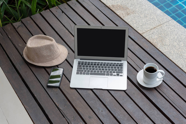 Laptop und Smartphone mit Kaffeetasse und Hut auf Holz. Laptop oder Notizbuch und Hut am Pool.  - Foto, Bild