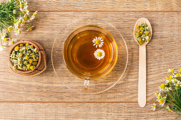 Стеклянная чашка зеленого чая с белыми ромашками и маленькая деревянная бочка с сухими цветами ромашки на досках. Вид сверху
 - Фото, изображение