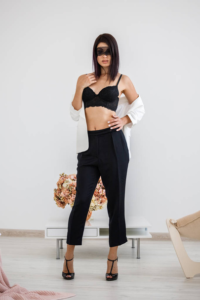 Studio portrait of beautiful brunette woman with black mask on face, wearing black pants, white jacket and black bra standing at home with flowers bouquet on background  - Photo, Image