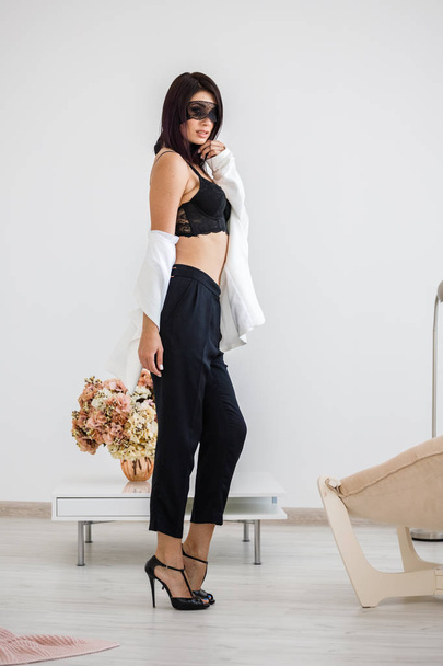 Studio portrait of beautiful brunette woman with black mask on face, wearing black pants, white jacket and black bra standing at home with flowers bouquet on background  - Foto, afbeelding
