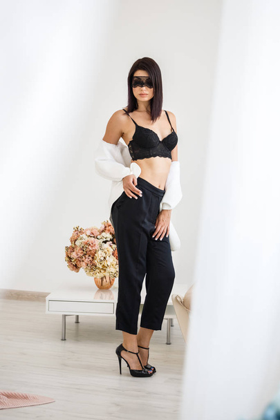 Studio portrait of beautiful brunette woman with black mask on face, wearing black pants, white jacket and black bra standing at home with flowers bouquet on background  - Photo, image