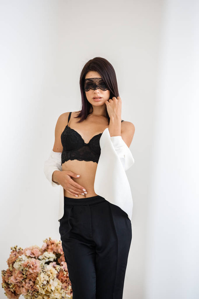 Studio portrait of beautiful brunette woman with black mask on face, wearing black pants, white jacket and black bra standing at home with flowers bouquet on background  - Fotó, kép