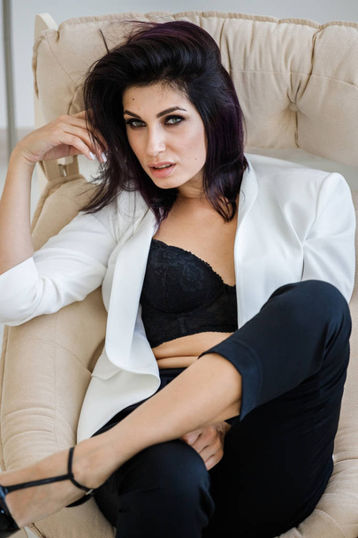 Studio portrait of beautiful brunette woman wearing black pants, white jacket and black bra lying in armchair at home interior  - Foto, immagini